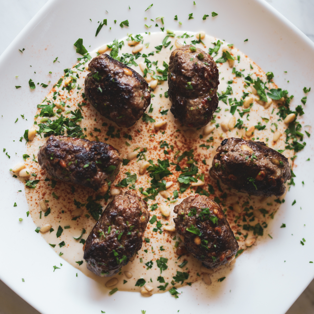 Kofta b'siniyah garnished with pine nuts and parsley on a white plate with a tahini sauce. Th