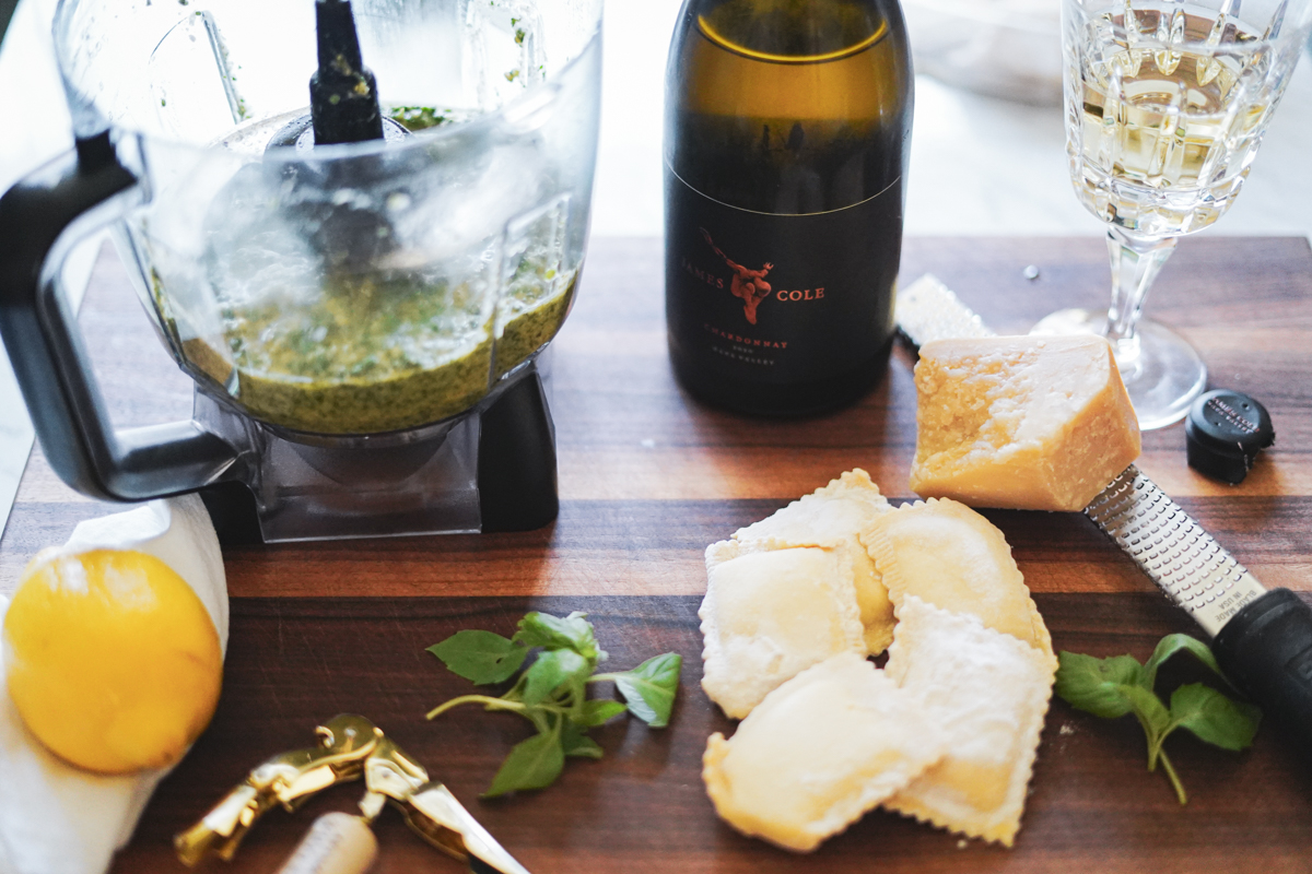 A bottle of James Cole 2020 Chardonnay on a cutting board with a container of Pesto a lemon, a few basil sprigs, ravioli and a microplane with a block of parmesan.