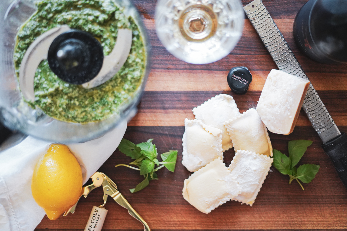 A wooden cutting board with a container of pesto, a glass of wine, a lemon, basil, ravioli, a microplane and block of parmesan.