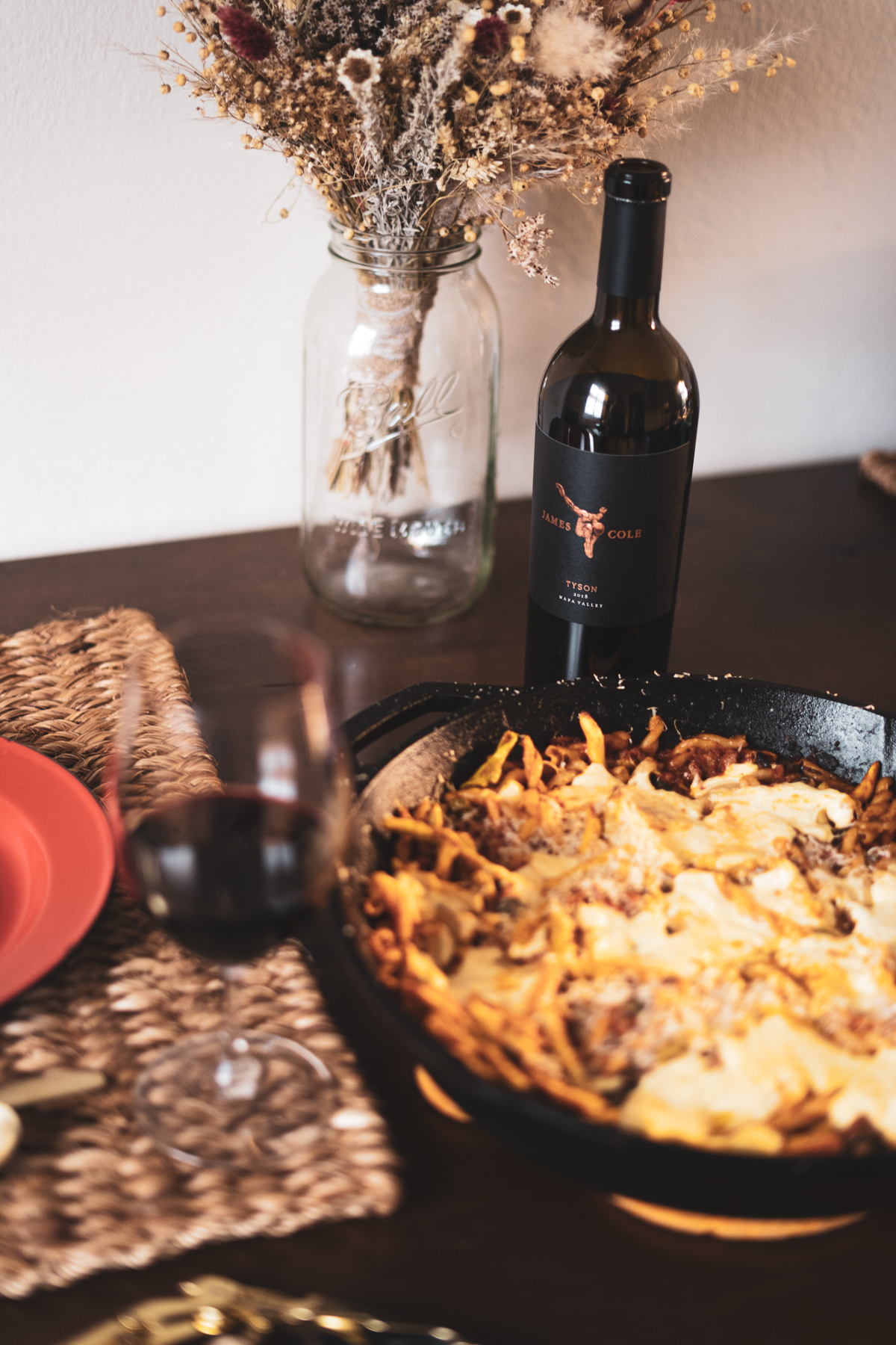 Baked Ziti paired with 2018 Tyson Red Wine. The dinner is set at a dark brown wooden table with jute placemats and a dried floral bouquet can be seen in the background.