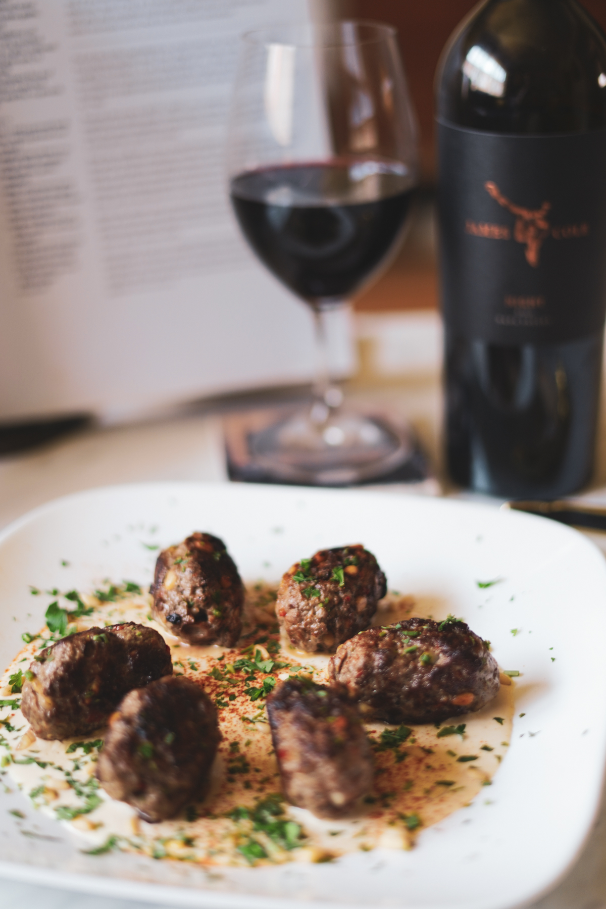 Lamb kofta served on a white plate with a bottle and glass of 2020 Night Red Wine in the background.